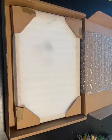 The box opened: the frame under a foam sheet, held by cardboard corners, beside an air pillow